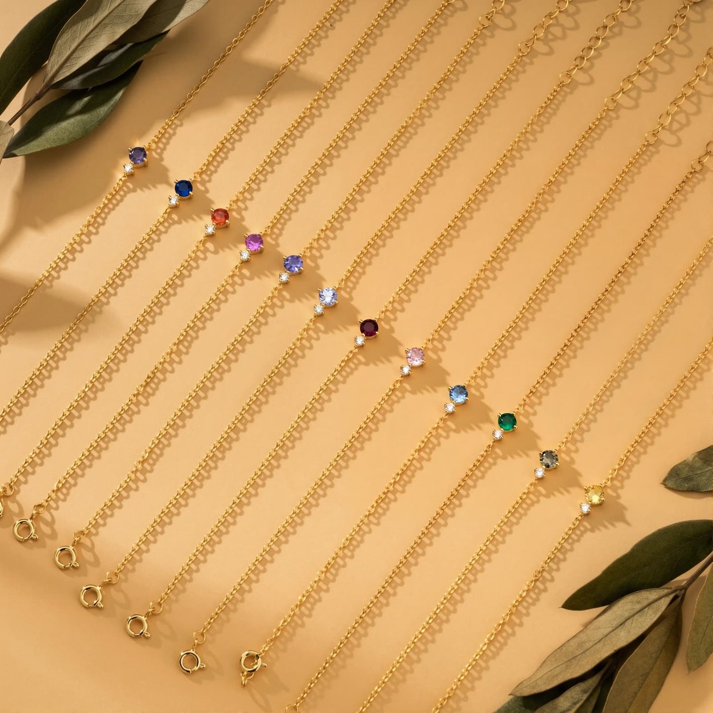 Twelve gold-tone birthstone bracelets in different colors arranged with leaves on a warm background