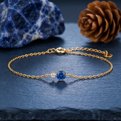 Gold-tone bracelet with a blue round birthstone and clear side accent on a dark stone surface