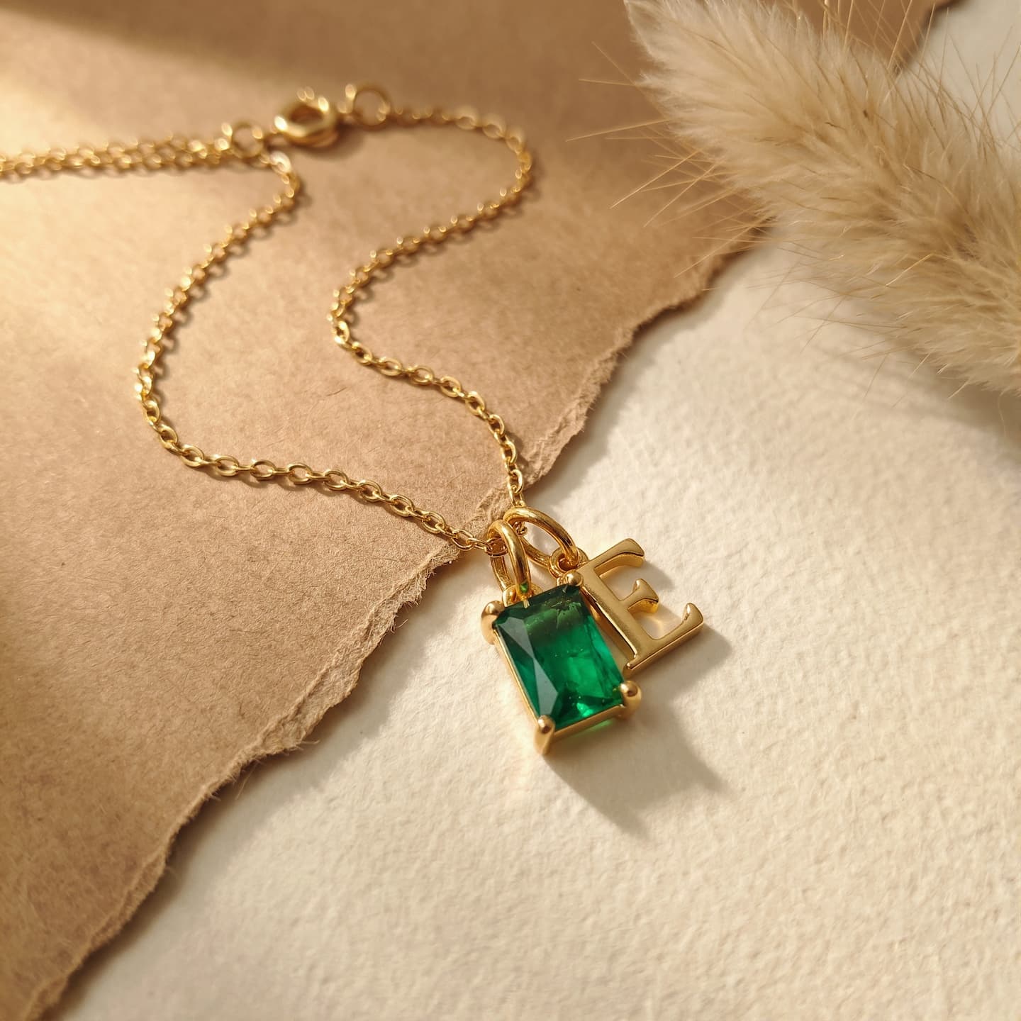 Gold-tone necklace with a green rectangular birthstone and uppercase E initial on layered beige and cream fabric