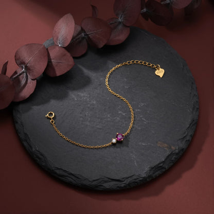 Gold-tone bracelet with a pink round birthstone, clear side accent, and spring-ring clasp on a dark round surface