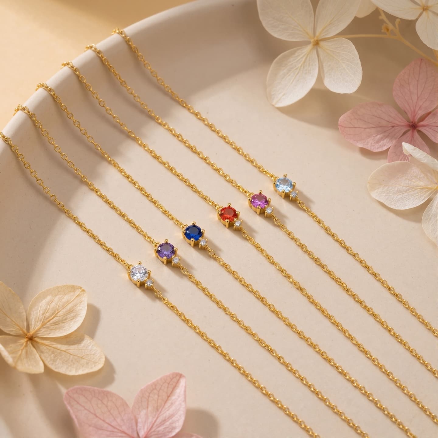 Five gold-tone birthstone bracelets in different colors arranged on a cream tray with pale flowers