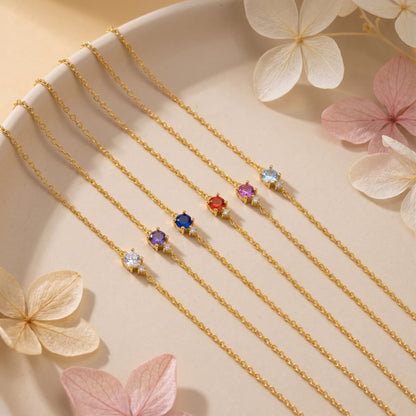 Five gold-tone birthstone bracelets in different colors arranged on a cream tray with pale flowers
