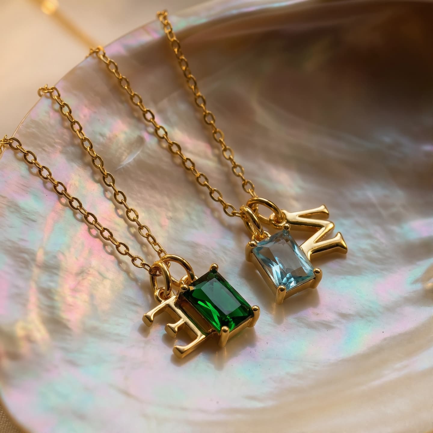 Two gold-tone birthstone necklaces with uppercase E and M initials arranged on an iridescent shell