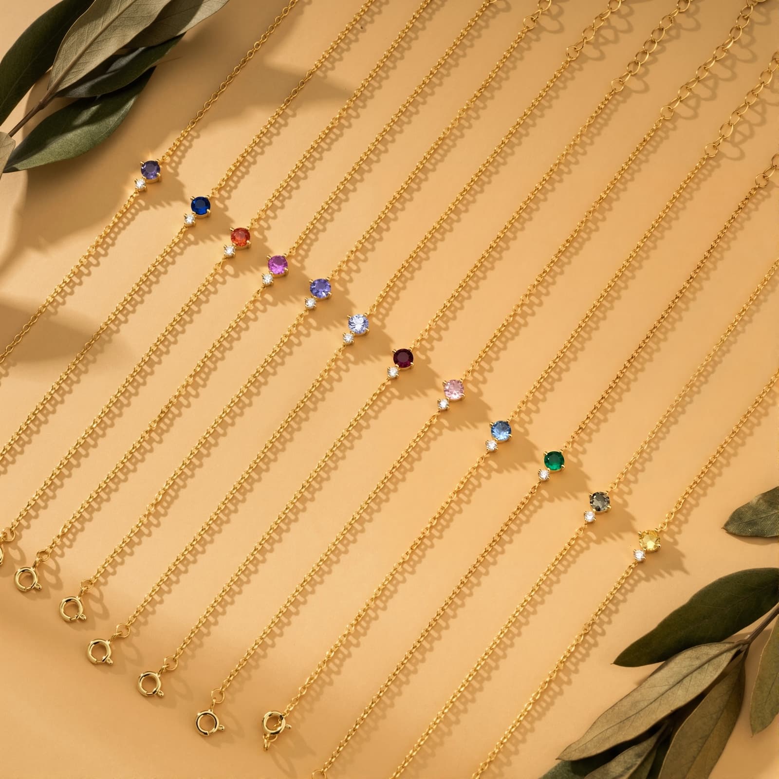 Twelve gold-tone birthstone bracelets in different colors arranged with leaves on a warm background