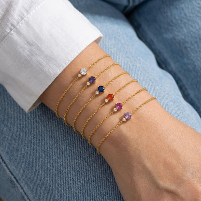 Six gold-tone birthstone bracelets in different colors stacked over a white sleeve and denim