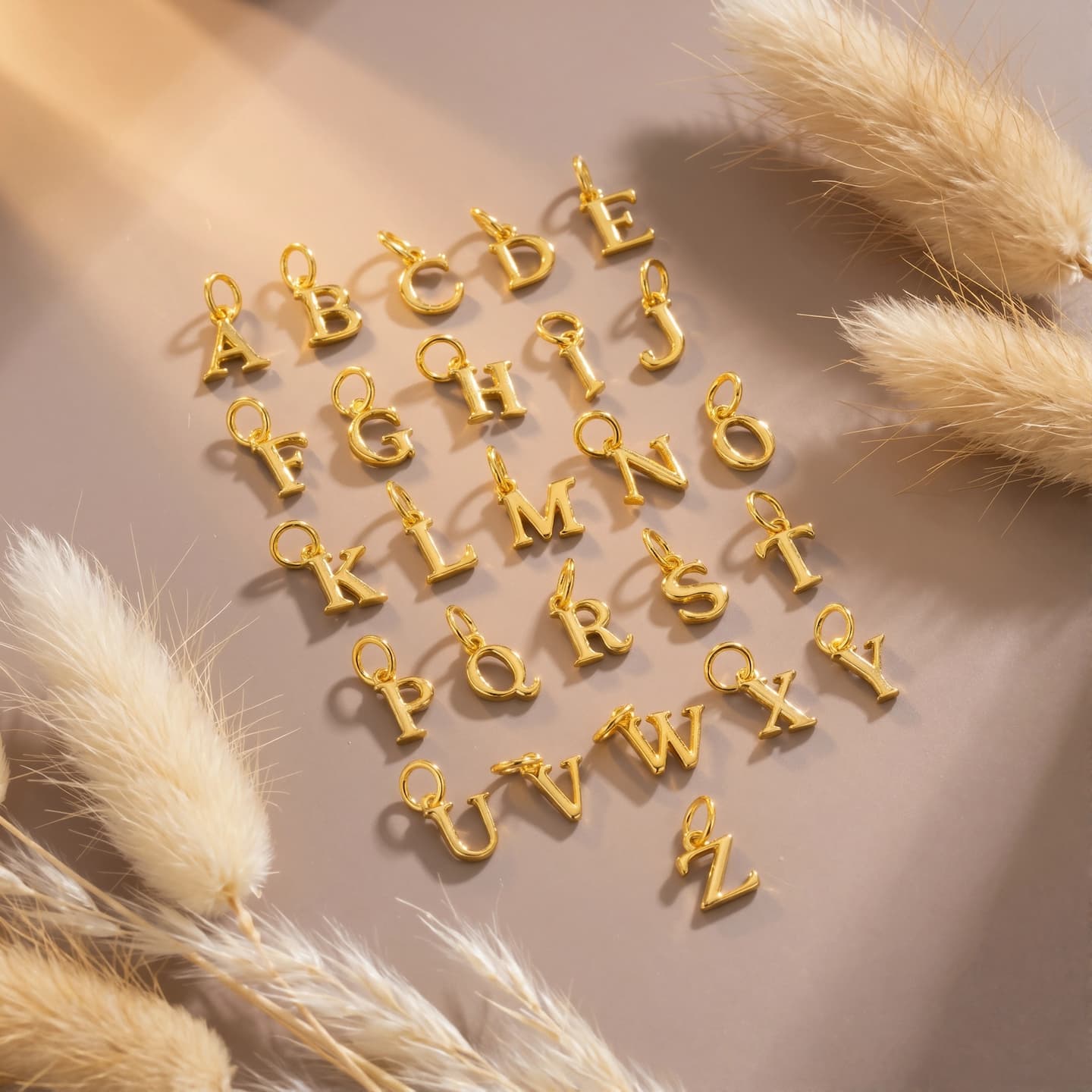 Gold-tone uppercase A-Z initial charms arranged on a warm neutral background with dried grasses