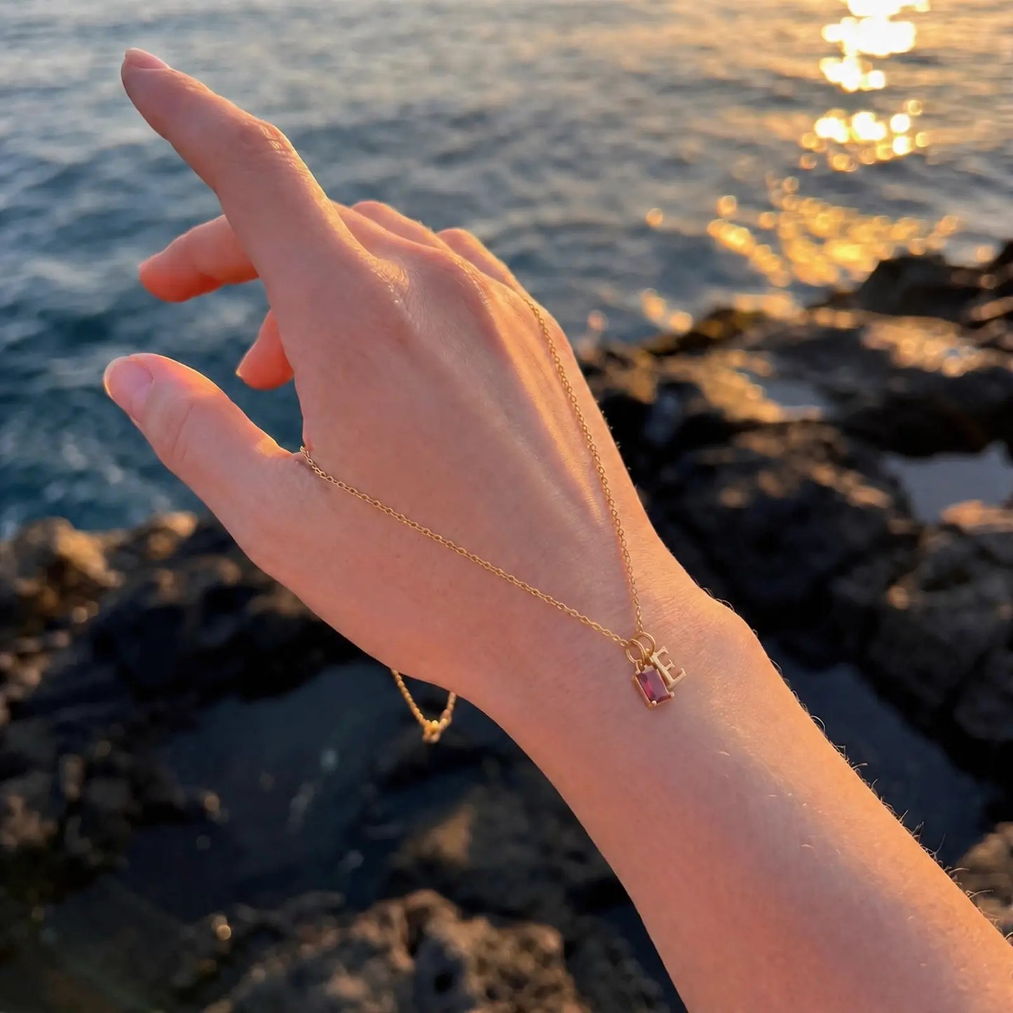 Hand holding a delicate birthstone and initial necklace beside the sea at sunset