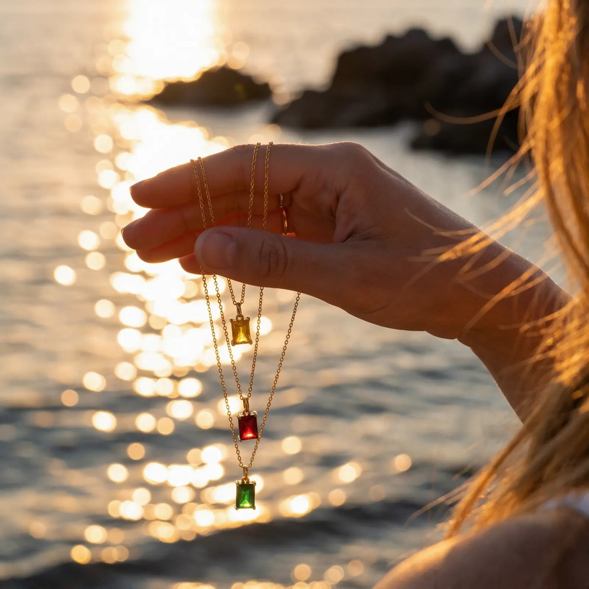 Three rectangular birthstone pendant necklaces held by the sea at sunset